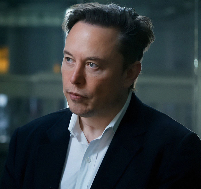 A man with short hair, in a suit, looking thoughtful in an indoor setting.