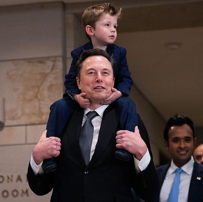 Man in a suit carrying a child on his shoulders indoors, with people in the background.