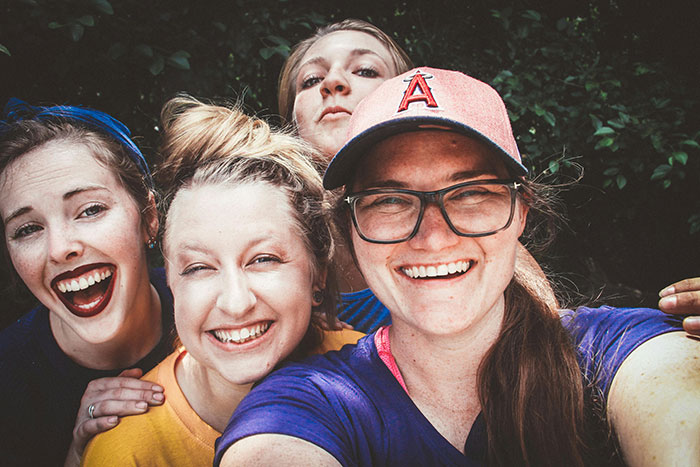 Four women smiling for a selfie outdoors, one wearing a cap and glasses.