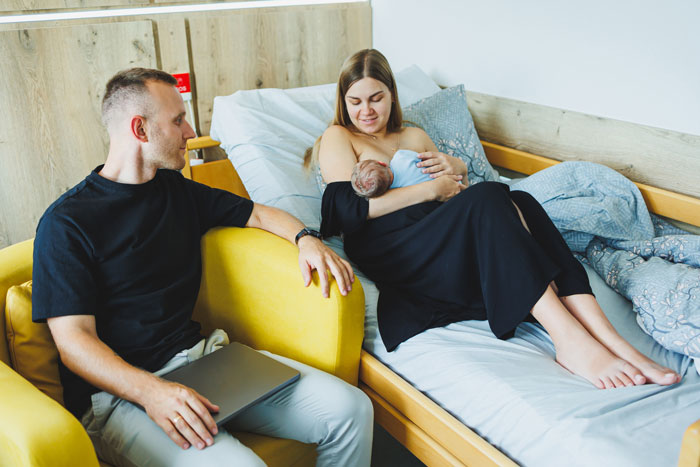 Woman with newborn baby in hospital bed, man sitting nearby holding a laptop, conveying family emotions after birth.