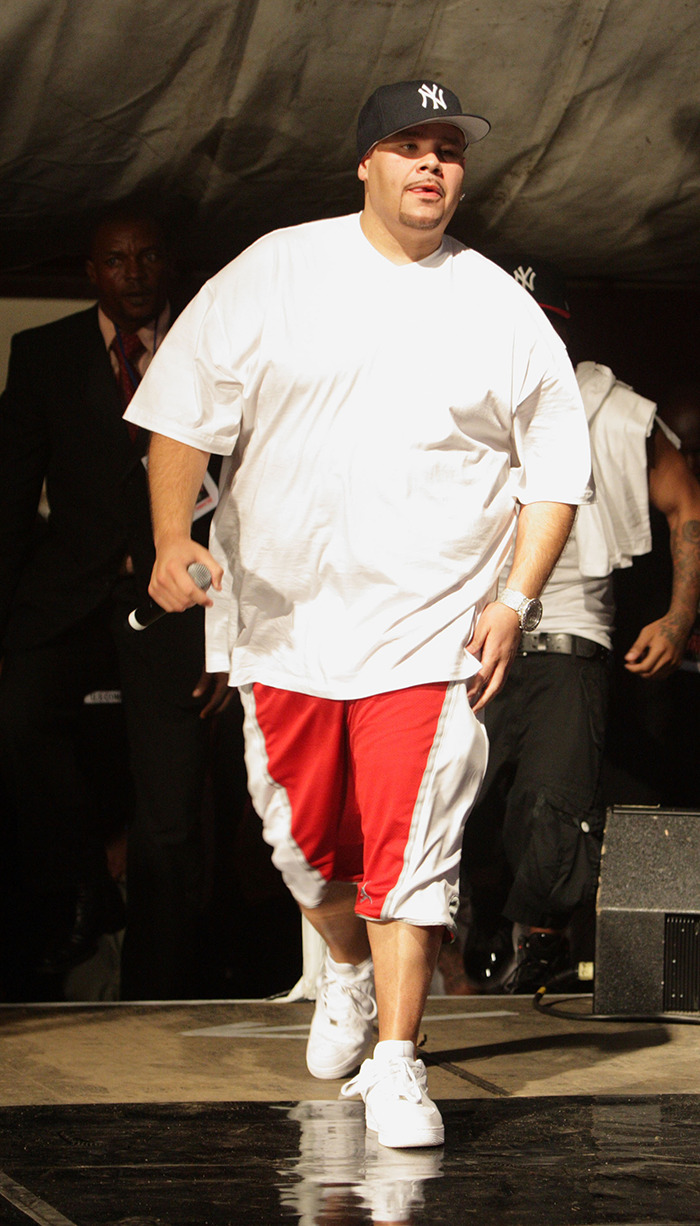 Man in oversized white shirt and red shorts walking on stage, holding a microphone; raising questions about beauty standards.