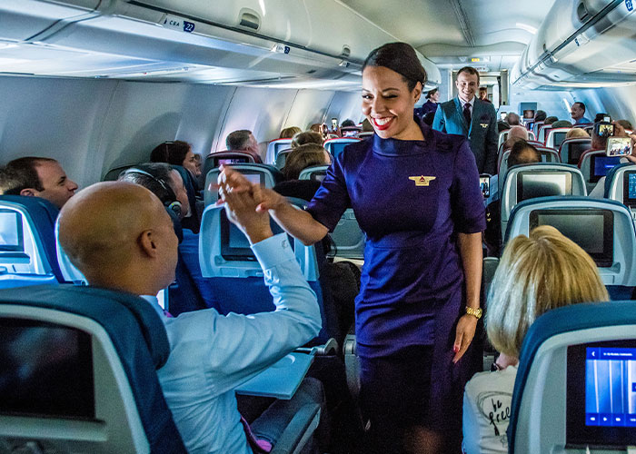 Flight attendant interacting with passengers in an airplane aisle, smiling and engaging in conversation.