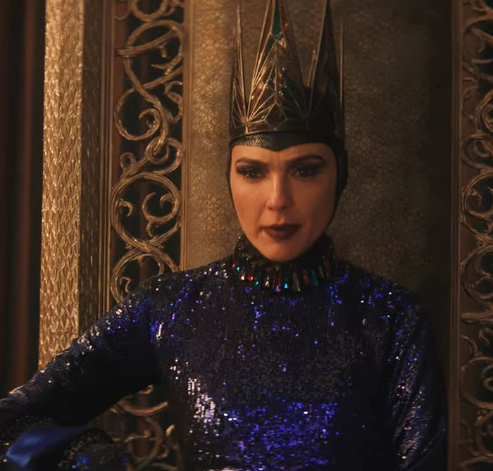 A woman in a detailed dark costume with a crown, part of the Disney 'Snow White' film, seated on an ornate chair.