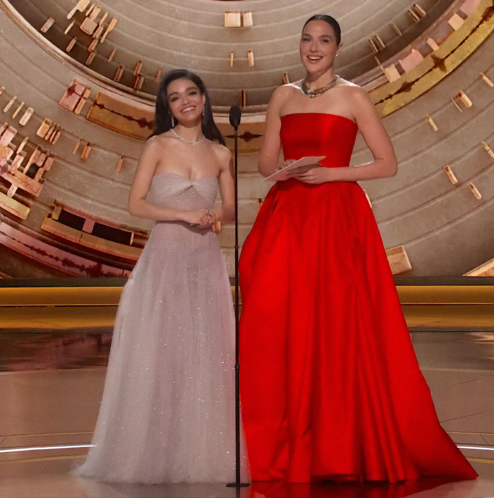 Two women elegantly dressed on stage at an event, one in a silver gown and the other in red.