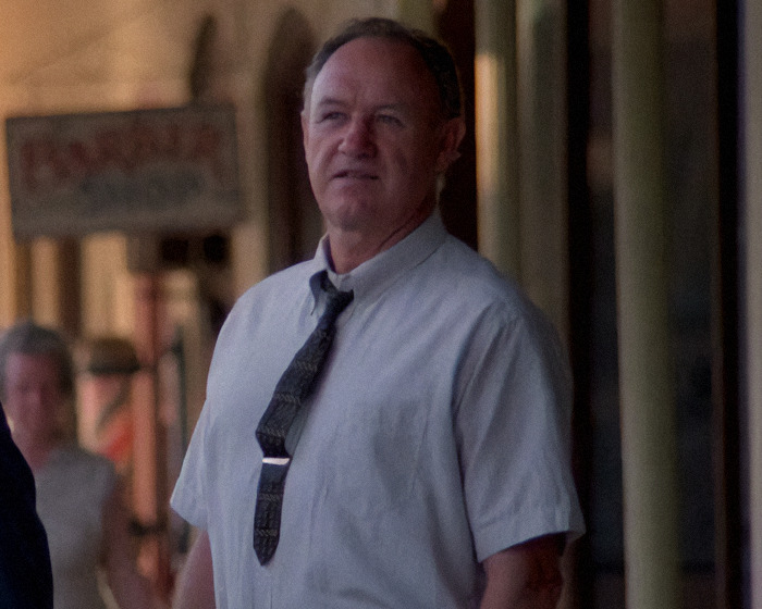 Gene Hackman on a street, wearing a tie and shirt, looks into the distance with a blurred backdrop of a shop sign.