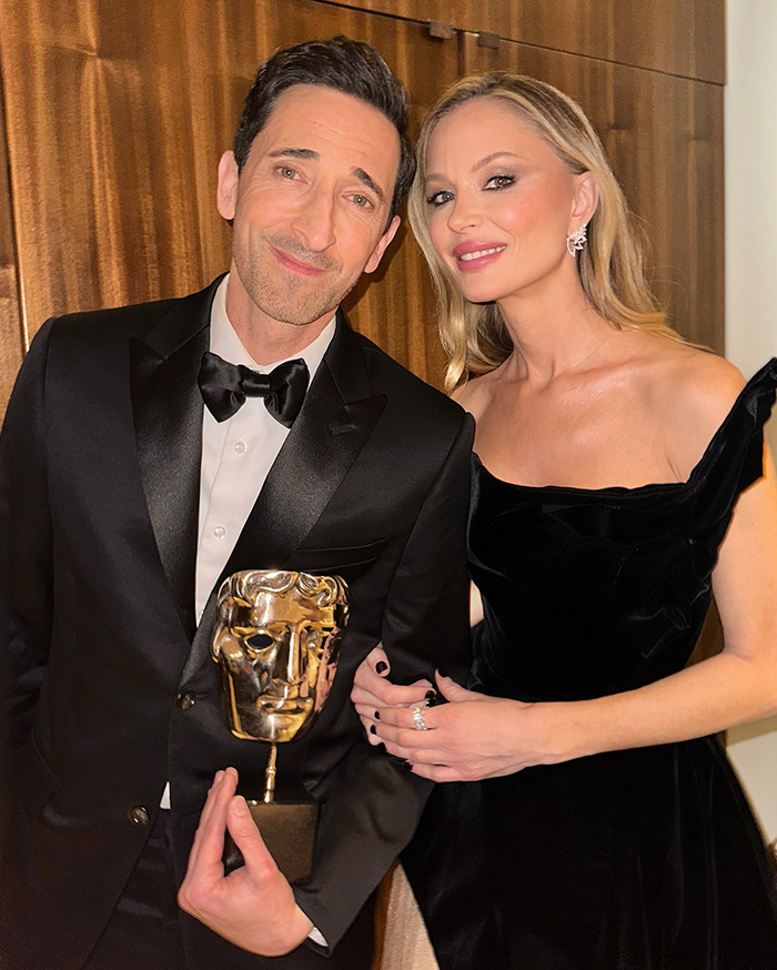 Adrien Brody holding award, smiling with a woman, after Oscars speech mentioning children, wearing black formal attire.