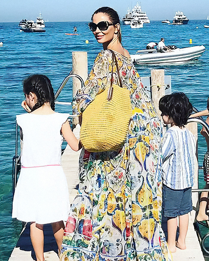 Georgina Chapman in a colorful dress and sunglasses, holding hands with two children on a pier, with boats in the background.