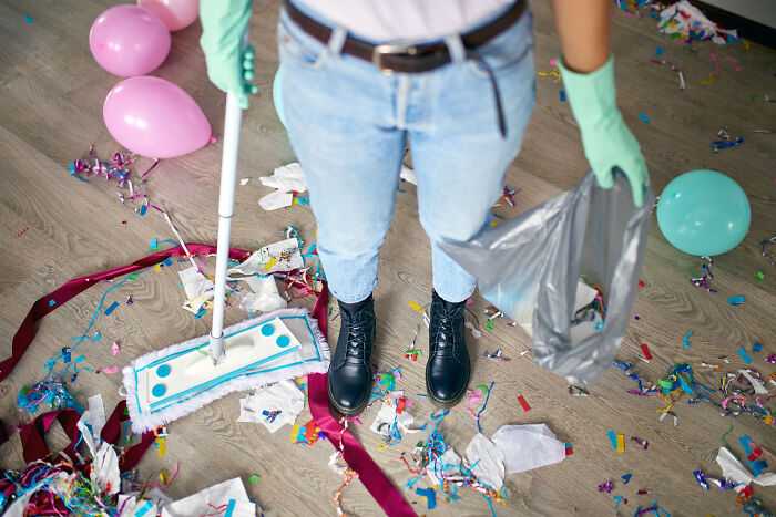 Person using cleaning hacks to tidy up colorful party mess with a mop and trash bag.