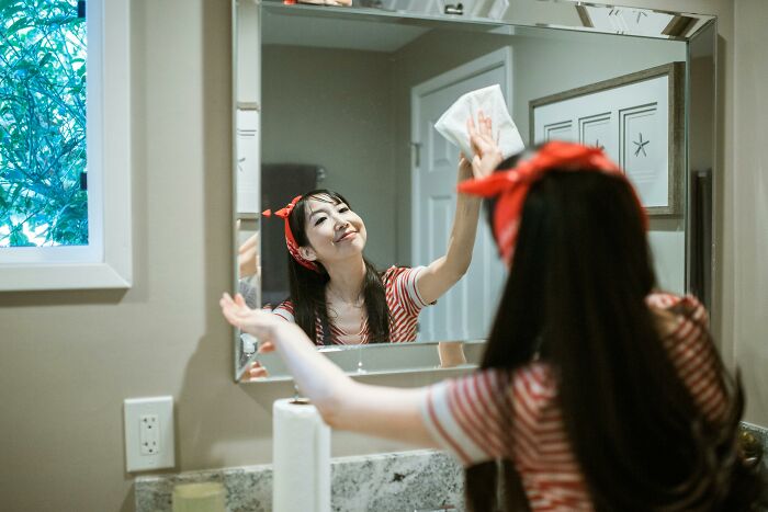 Woman using cleaning hack in a bathroom, wiping a mirror with a cloth, wearing red headband and striped shirt.