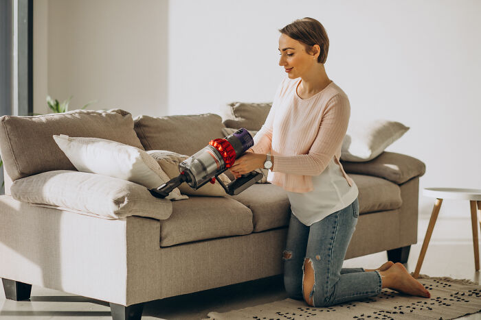 Woman using a vacuum cleaner on a sofa, demonstrating a lesser-known cleaning hack in a bright, cozy living room.