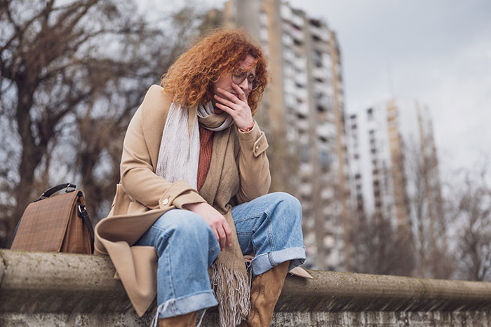 Woman with red curly hair sitting outdoors, looking pensive and upset, with urban background; theme of having 1 child.