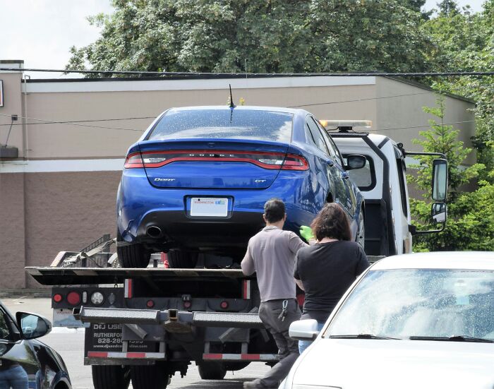 Blue Dodge Dart GT being loaded onto a tow truck in a parking lot with two people overseeing the process.