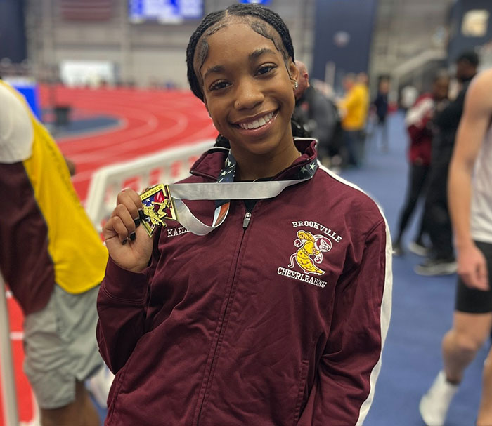 Smiling track star holding a medal in a maroon jacket at an indoor track event.