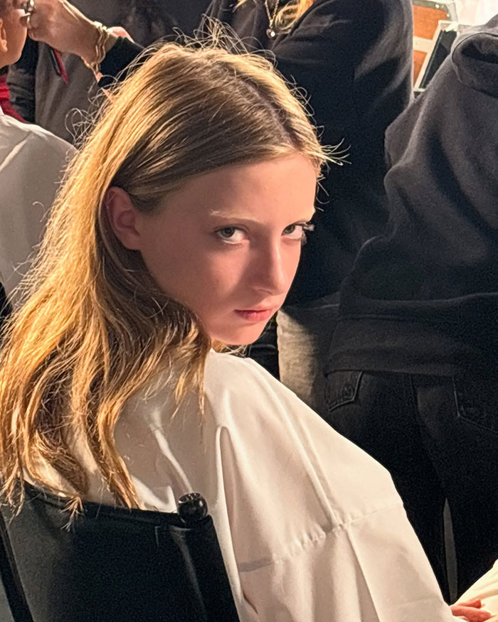 Young Model en la Semana de la Moda de París, sentado en una silla, con un atuendo blanco, peinado, rodeado de personas en el backstage.