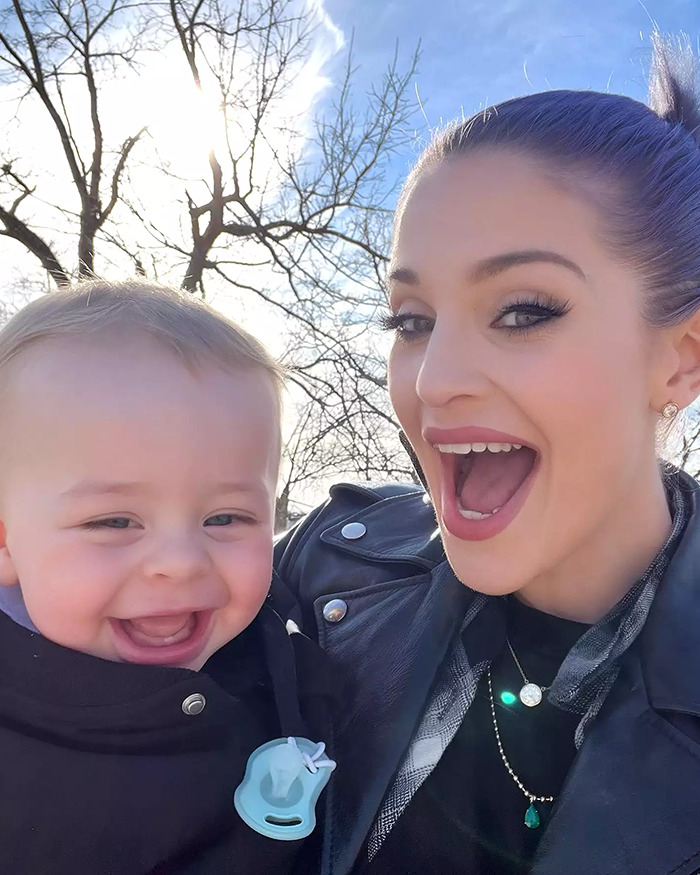 Woman with dramatic weight loss smiling outside, wearing a black leather jacket, holding a baby in winter attire.