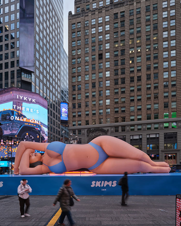 Giant bikini blow-up doll in Times Square causes backlash, surrounded by skyscrapers and pedestrians.