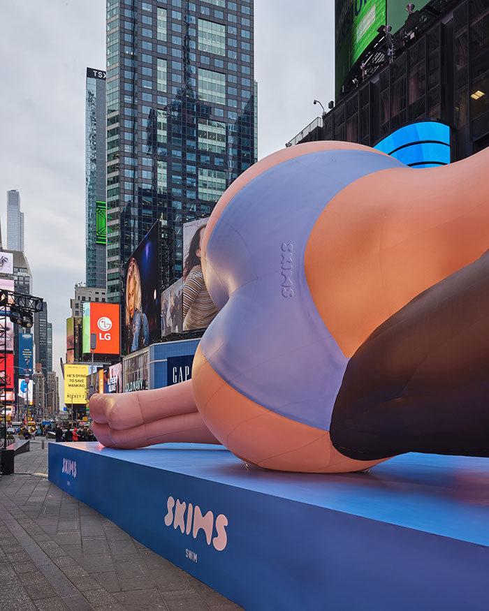 Giant bikini blow-up doll in Times Square promoting Skims brand, surrounded by skyscrapers and billboards.