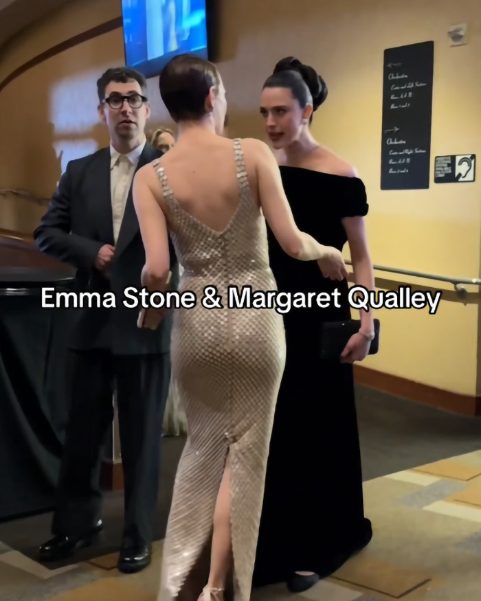 Emma Stone and Margaret Qualley in an animated exchange at an event, attended by a suited gentleman in the background.