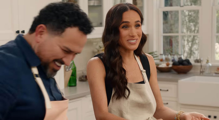 Two people in a kitchen, one wearing an apron, discussing Prince Harry.