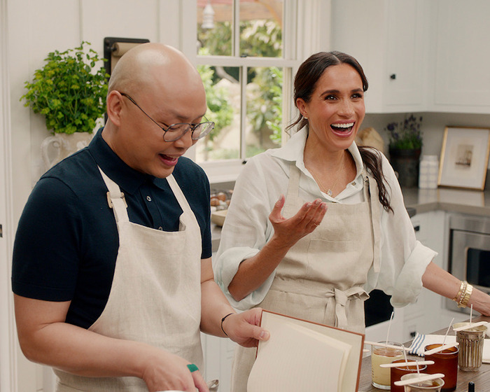 Two people smiling in a kitchen, wearing aprons, discussing a project related to Meghan Markle's show controversies.