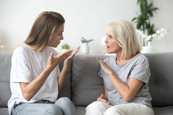 Daughter-in-law and MIL in a heated discussion on a gray couch, both gesturing expressively.