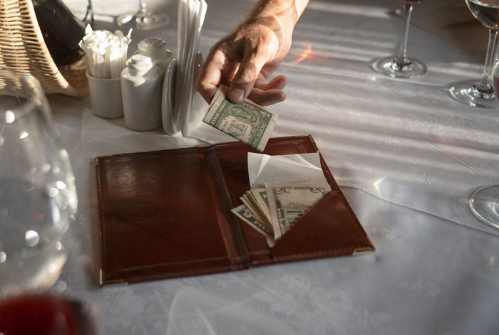 Hand placing cash in a brown bill holder on a restaurant table, highlighting wallet absence.