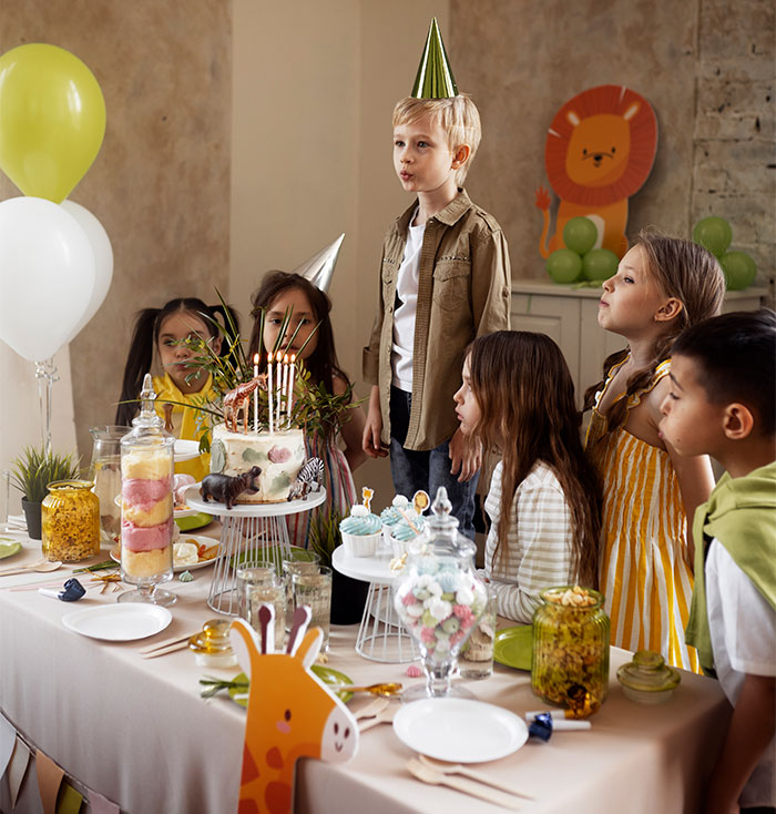 Children at a birthday party looking at a cake, wearing party hats, and surrounded by balloons and decorations.