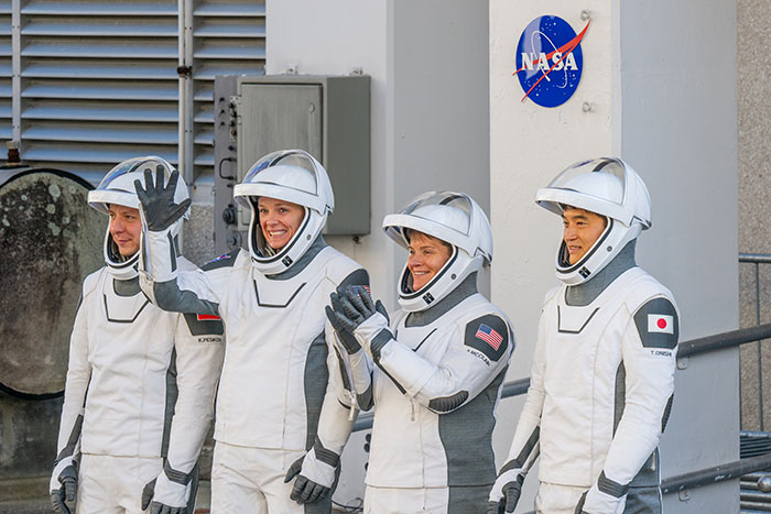 Astronauts in SpaceX suits smiling and waving near NASA emblem, involved in delayed rescue mission.