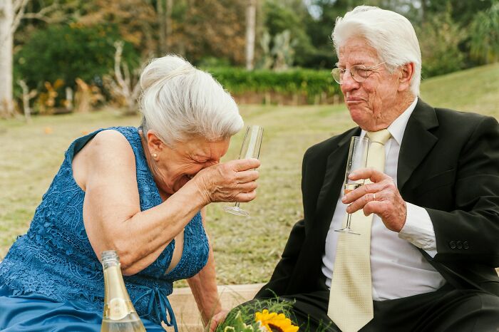 Elderly couple in formal attire, sitting outdoors, sharing a laugh with champagne during a wedding objection story moment.