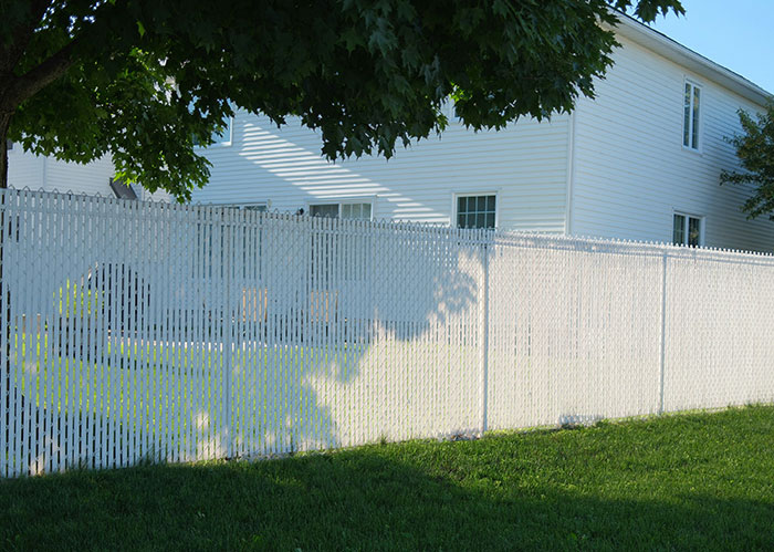 White fence casting shadows on grass, separating houses in a neighborhood with green trees and blue skies.