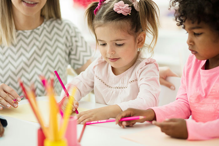 Young kids coloring with pencils at a daycare table, guided by a caregiver.