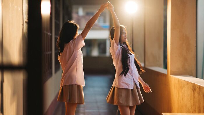 Two girls dancing in a sunlit corridor, dressed in uniforms, with warm light creating a serene world perspective.