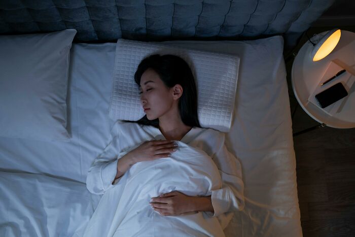 Woman sleeping comfortably in bed, with a lamp and phone on the nightstand.