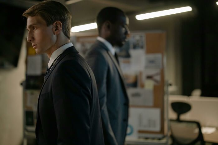 Two men in suits standing in an office, with Wikipedia articles on a bulletin board behind them.