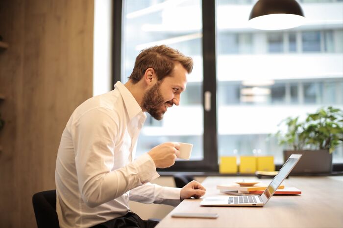 Man with a beard sitting at a desk, smiling at a laptop, holding a coffee cup.