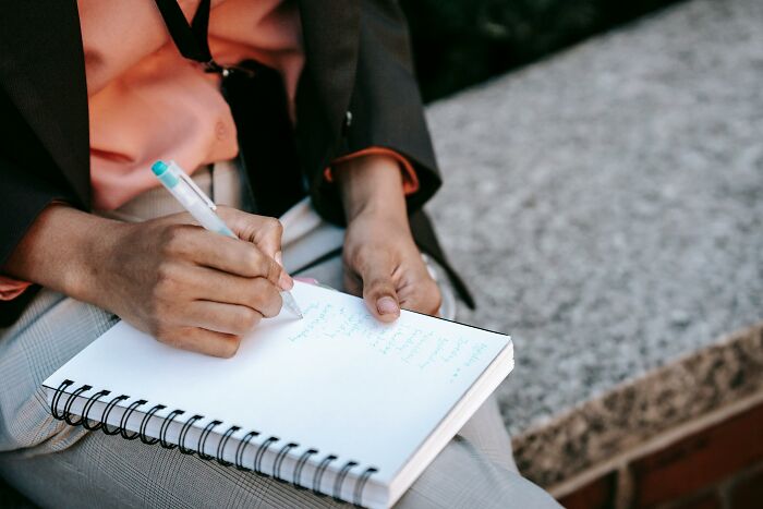 Person writing in a notebook outdoors, possibly reading cool Wikipedia articles, with a pen in hand and wearing a blazer.