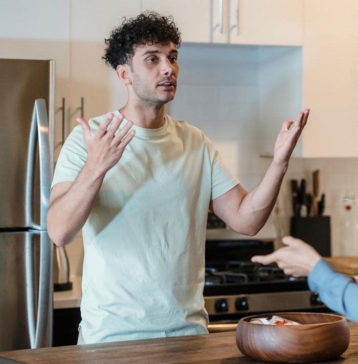 Man discussing in kitchen while gesturing, partner&rsquo;s hand pointing back, highlighting relationship and sterilization topic.