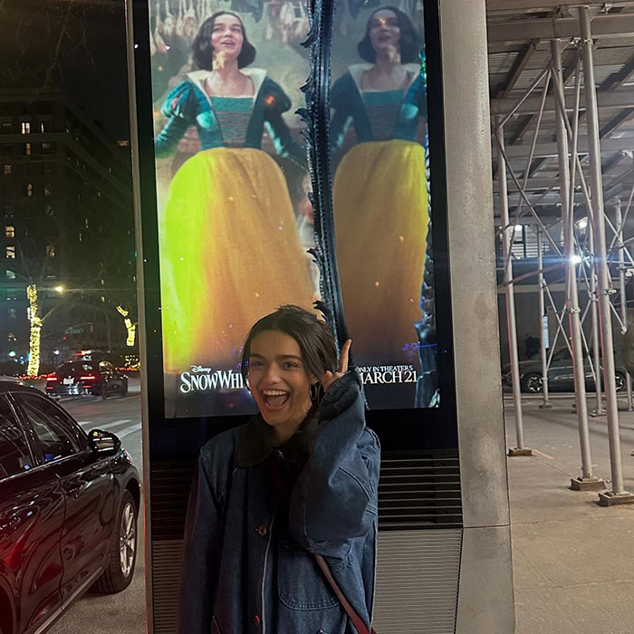 A woman smiles beside a digital poster for Disney's 'Snow White' at night, with city lights in the background.