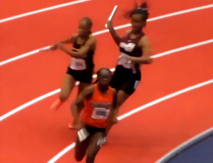 Track star in a relay race, caught in action with a baton, surrounded by competitors on a red track.