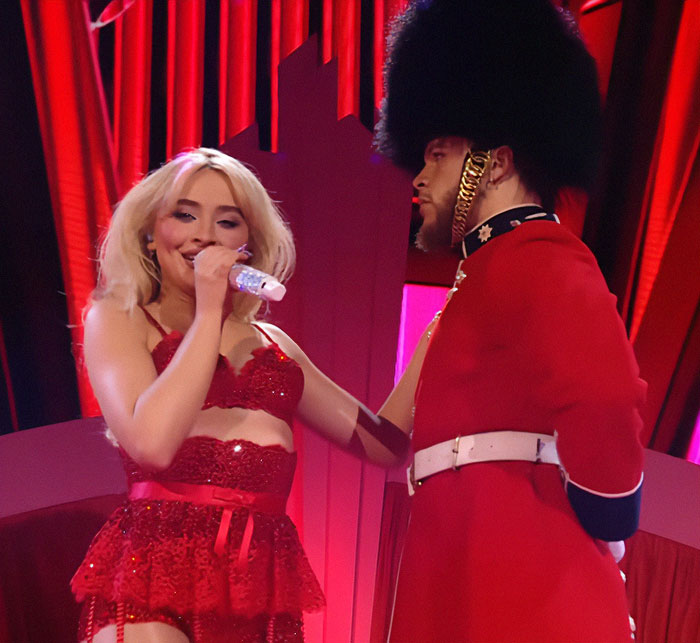 Singer performing in red outfit with guard in themed concert scene.