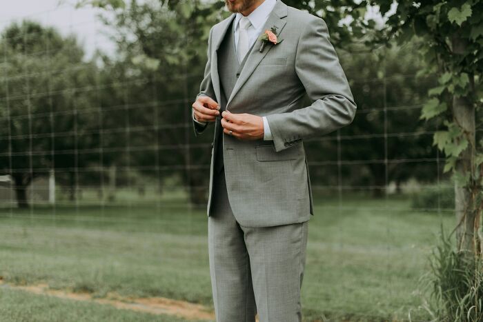 Groom in gray suit adjusts jacket outdoors, wedding setting.