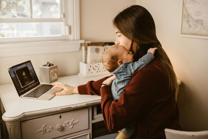 A woman holding a baby while using a laptop, representing the challenges of babysitting duties.