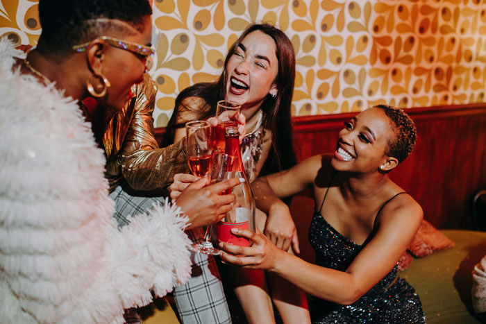 Three women celebrating with drinks, laughing together at a cozy gathering, highlighting babysit and family dynamics.