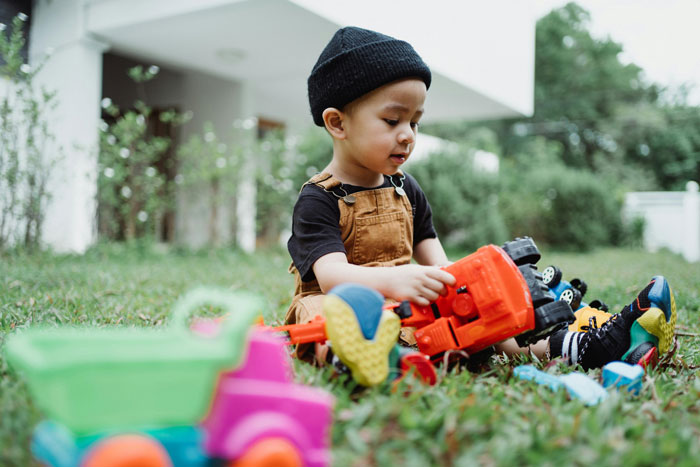 Child playing with toys outside on grass, wearing overalls and a beanie.