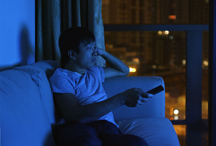 Man in a dark room, sitting on a couch watching TV, highlighting a struggle with his living situation and rules.