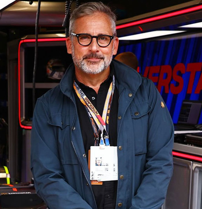 Man in a navy jacket smiling, standing indoors, wearing glasses, with lanyards around his neck.