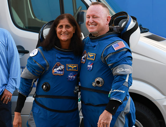 Astronauts in blue suits smiling, emblem patches visible, standing outside a vehicle.