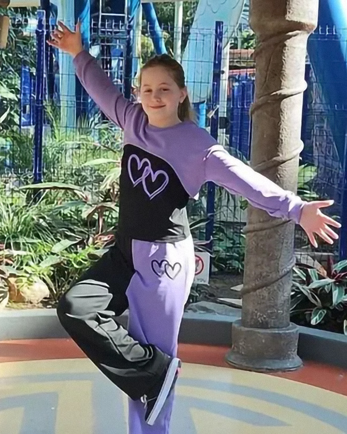 Young girl in purple outfit posing with arms outstretched at a park, near plants and a tree pillar.