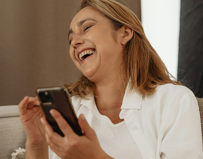 Woman in white shirt laughing while looking at her phone, related to Tesla driver experiences at charging stations.