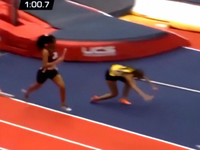 Track star with baton running alongside opponent on an indoor track.